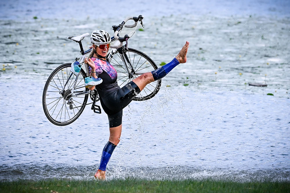 Patrycja Adaszewska z rowerem na plecach, krocząca po brzegu wody.